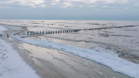 Aerial-establishing-view-of-an-old-wooden-pier-at-the-Baltic-sea-coastline,-overcast-winter-day,-white-sand-beach-covered-in-snow,-ice-on-wood-poles,-calm-seashore,-wide-drone-shot-moving-forward-low