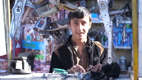 Junger-Mann-Am-Verkaufsstand-Im-Freien-Mit-Hängenden-Autoteilen,-Die-In-Quetta-In-Die-Kamera-Blicken