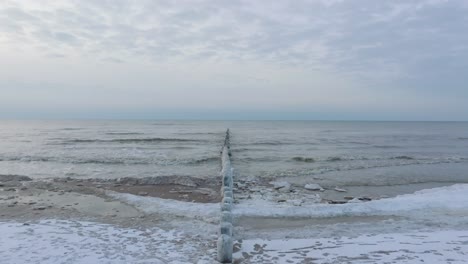 Aerial-establishing-view-of-an-old-wooden-pier-at-the-Baltic-sea-coastline,-overcast-winter-day,-white-sand-beach-covered-in-snow,-ice-on-wood-poles,-calm-seashore,-wide-drone-shot-moving-forward-low