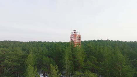 Aerial-establishing-view-of-Bernati-lighthouse-surrounded-by-lush-green-pine-tree-forest-with-light-snow,-Nordic-woodland,-Baltic-sea-coast,-sunny-winter-day,-Latvia,-wide-ascending-drone-shot