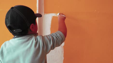 Kind-Trägt-Mütze-Mit-Malpinsel,-Um-Orangefarbene-Wand-In-Weiße-Farbe-Zu-Streichen