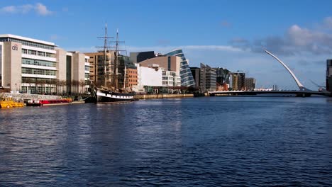An-over-water-slow-zoom-shot-of-the-River-Liffey-in-the-evening-sunshine,-showing-the-North-Wall-Quay-with-the-Samuel-Beckett-Bridge-in-the-distance