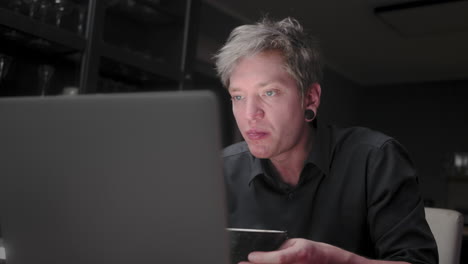 Young-man-with-silver-hair-and-a-black-business-shirt-sitting-lonely-in-a-dark-room-eating-asian-noodle-soup-andwiping-his-mouth,-while-staring-into-a-bright-notebook-screen