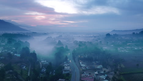 Wide-aerial-footage-of-the-road-and-cars-traveling-in-the-blue-light-of-the-sunrise-of-the-city-of-San-Juan-Ostuncalco-and-the-mountains-behind-it-in-the-haze
