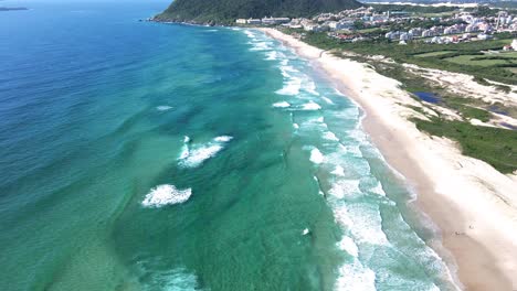 Aerial-drone-scene-of-paradisiacal-beach-with-turquoise-blue-and-emerald-green-ocean-with-mountains-and-vacation-resort-for-tourists-facing-the-beach-in-summer-in-florianópolis-Santinho-beach