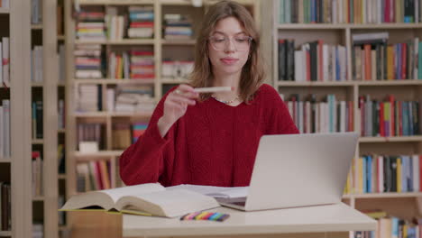 Young-woman-in-library-researching-with-laptop-and-books