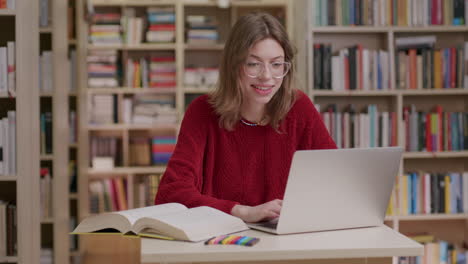 Smiling-caucasian-lady-wearing-red-sweater-and-glasses-work-on-laptop-in-library