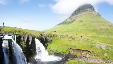 Kirkjufell,-and-Kirkjufellfoss-waterfall-in-Iceland,-with-people-walking-atop-the-waterfall-on-a-blue-sky-sunny-day-and-a-cloud-over-the-mountain