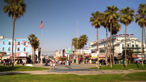 Panorámica-En-4k-De-Venice-Beach-En-Los-Ángeles,-Parque-Con-Césped-Verde,-Palmeras,-Edificios-Y-Bandera-De-Estados-Unidos-En-El-Fondo,-Jóvenes-Caminando-Y-Scooters