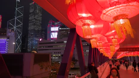 Menschen-Gehen-Durch-Eine-Fußgängerbrücke,-Die-Mit-Chinesischen-Laternen-Geschmückt-Ist,-Die-Von-Der-Decke-Hängen,-Um-Das-Chinesische-Neujahrsfest-Zu-Feiern,-Während-Die-Skyline-Von-Hongkong-Auf-Der-Linken-Seite-Zu-Sehen-Ist