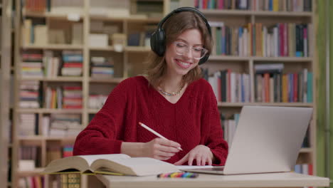 Niña-Sonriente-Con-Gafas-Y-Auriculares-Estudia-Con-Una-Laptop-En-La-Biblioteca