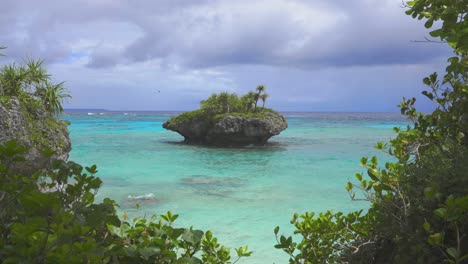 A-small-rocky-island,-with-its-rugged-and-textured-surface,-sits-majestically-in-front-of-a-tropical-beach,-surrounded-by-crystal-clear-waters