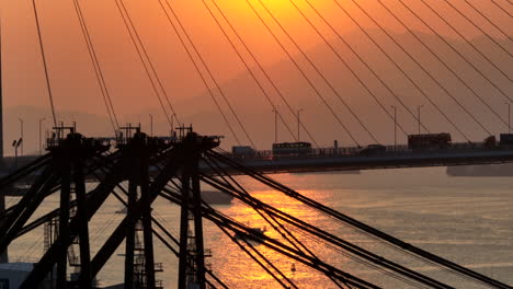 Aerial-descending-shot-of-suspension-bridge-with-traffic-and-container-terminal-silhouette-with-golden-sunset-in-background