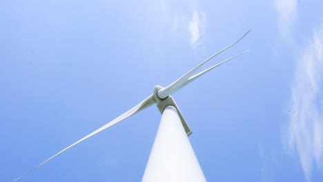 Low-angle-looking-up-at-wind-turbine-spinning-blades-renewable-energy-generator-against-blue-sky