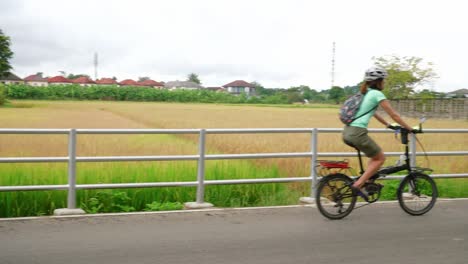 Mujer-Asiática-Montando-Lentamente-En-Bicicleta-Por-Una-Carretera-Pavimentada-En-Una-Ciudad-Rural-Durante-El-Día,-Tailandia