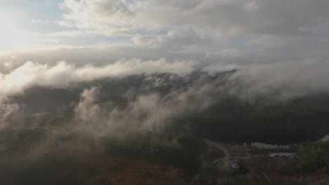 Drone-footage-of-highway,-and-housing-estate-in-Norway-with-some-fog-floating-in-the-air-and-sun-lighting-up-the-clouds