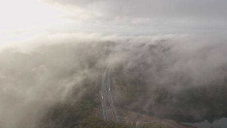 Drone-footage-of-highway,-and-housing-estate-in-Norway-with-some-fog-floating-in-the-air-and-sun-lighting-up-the-clouds