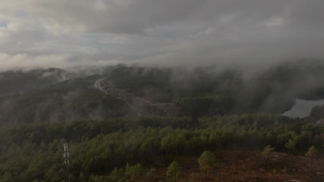 Drone-footage-of-highway,-radiotower,-and-housing-estate-in-Norway-with-some-fog-floating-in-the-air-and-sun-lighting-up-the-clouds