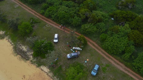A-drone-captures-a-shot-of-people-enjoying-a-picnic-near-a-dirt-road,-showcasing-the-natural-scenery-surrounding-them,-including-the-sandy-beach-and-the-lush-greenery-of-the-trees-during-the-day