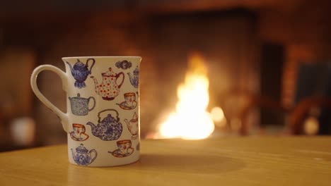 A-white-coffee-mug-sitting-by-a-warm-fire-on-a-wooden-table-inside-a-house-on-a-cold-winter-night