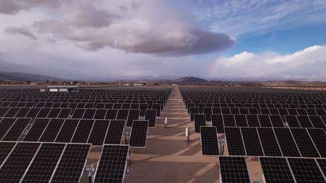 Aerial-Drone-Footage-of-Solar-Panel-Field-in-Joshua-Tree-National-Park-on-a-Sunny-Day-with-rainbow-in-the-background,-fast-movement-forward-and-up