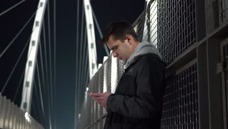 Young-male-teen-standing-while-on-his-phone-on-a-walking-bridge-at-night