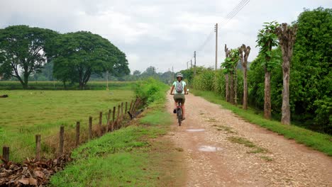 Female-riding-bike-on-rocky-dirt-path-in-countryside-away-from-camera,-Thailand
