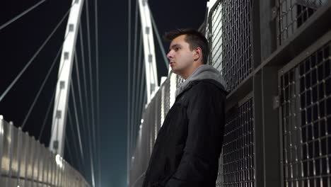 Young-white-male-teen-standing-on-a-bridge-at-night-looking-around