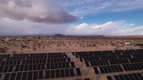 Aerial-Drone-Footage-of-Solar-Panel-Field-in-Joshua-Tree-National-Park-on-a-Sunny-Day-with-rainbow-in-the-background,-reveal-moving-backwards