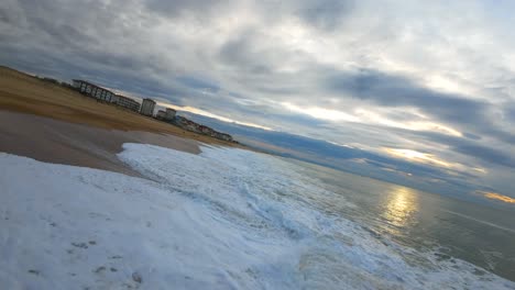 first-person-view-flight-over-the-waves-at-the-wild-surf-beach-of-Hossegor-during-a-cloudy-sunset