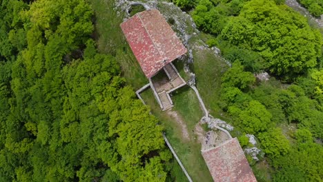 Drone-aerial-shot-captures-a-stone-hermitage-with-a-red-roof-hidden-among-the-lush-green-foliage-of-the-forest-on-the-peak-of-Mount-Erga