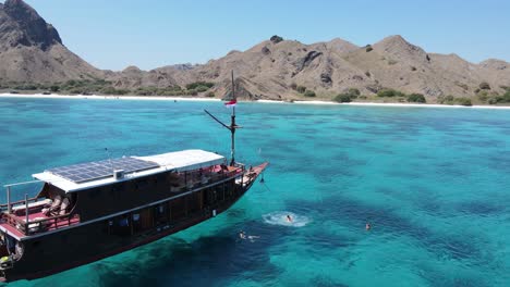 Aerial-View,-People-Jumping-in-Turquoise-Sea-Water-From-Ship-Anchored-by-Komodo-Island-Coast,-Indonesia
