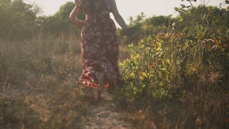 An-excited-Caucasian-female-wearing-a-red-floral-dress,-running-barefoot-towards-the-golden-sunset-on-a-beautiful-day-outdoors