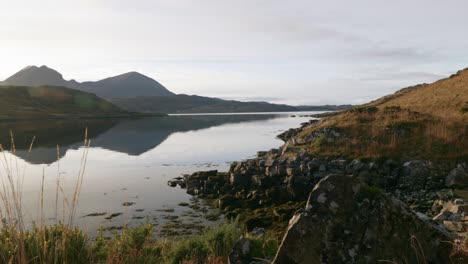 The-camera-gently-rises-above-vegetation-to-reveal-the-sun-reflecting-off-of-the-surface-of-a-tidal-sea-loch-in-the-north-west-Highlands-of-Scotland-just-before-setting-behind-mountains