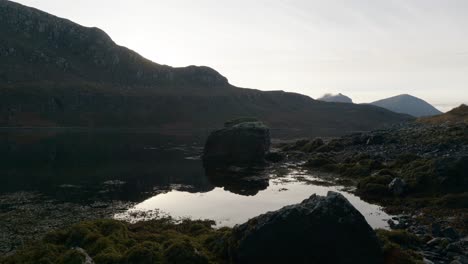 Gentle-ripples-move-across-the-surface-of-dark,-still-water,-tidal-pools-and-seaweed-in-a-sea-loch-in-Scotland-as-the-sun-sets-behind-mountains-