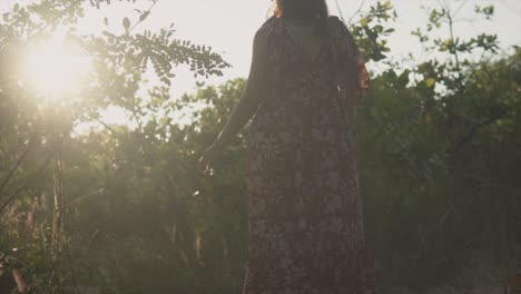 Low-angle-shot-of-the-back-of-a-Caucasian-female-walking-through-a-field,-the-golden-rays-of-the-distant-sunset-shining-through-the-gaps-of-the-trees-on-a-beautiful-carefree-day
