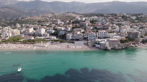 Aerial-view-of-Waterfront-Himare-Buildings-by-turquoise-colored-water,-Mountains-in-Background,-Albania