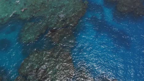 A-drone-captures-a-shot-of-a-blue-lagoon,-showcasing-the-crystal-clear-waters,-as-the-camera-pans-up-to-reveal-the-lush-coastal-tropical-landscape-with-a-distant-rainy-cloud
