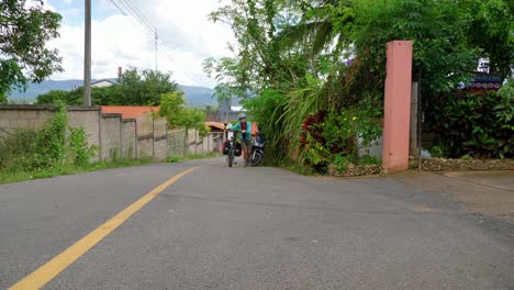 Vista-Estática-De-Una-Joven-Mochilera-Asiática-Caminando-Con-Su-Bicicleta-Por-La-Carretera-Rural-Del-Sudeste-Asiático