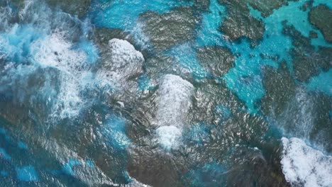 A-drone-captures-a-shot-of-a-blue-lagoon,-showcasing-the-clear-waters-that-reveal-the-underwater-landscape-of-rocks,-the-waves-crashing-on-the-reef-adding-to-the-dynamic-of-the-scene