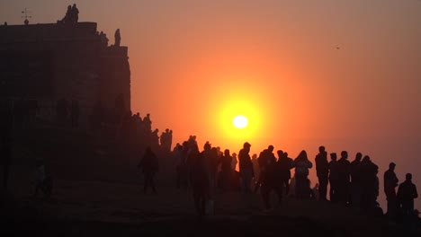 People-Silhouette-watching-surfers-riding-giant-waves-in-Nazare-during-Epic-Sunset,-Famous-big-waves-in-the-World