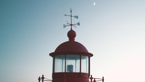 Old-red-and-charming-"Farol"-lighthouse-with-arrows-for-cardinal-points-and-moon-in-the-background-in-the-dark-sky,-Nazare-in-Portugal-late-summer-evening