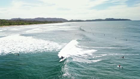 Luftaufnahme-Von-Vielen-Leuten,-Die-An-Einem-Sonnigen-Tag-In-Zeitlupe-Am-Strand-Von-Nosara-In-Costa-Rica-Surfen