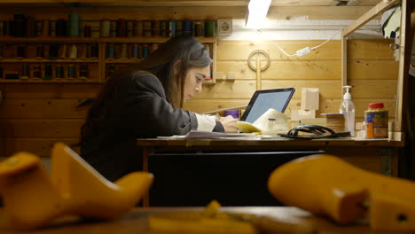 Female-craftsperson-drawing-shoe-design-artwork-under-spotlight-in-wood-panel-workshop