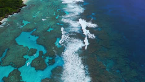A-drone-captures-a-shot-of-a-blue-lagoon-reef-with-waves-crashing,-displaying-the-vibrant-colors-and-movement-of-the-water,-panning-up-to-reveal-the-stunning-coastal-landscape-of-a-tropical-island