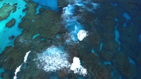 A-drone-captures-a-mesmerizing-shot-of-a-blue-lagoon-reef-with-waves-crashing-during-the-day,-showcasing-the-reef's-vast-beauty-and-the-dynamic-element-of-the-waves