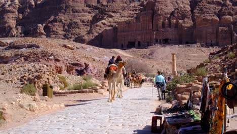 Arabian-people-riding-camels-through-the-desert-city-of-Petra-in-Jordan,-Middle-East,-with-tourist-wandering-and-exploring-the-Jordanian-ancient-city-tourism-landmark