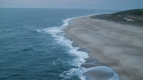 Große-Wellen-Brechen-An-Der-Küste-In-Nazare,-Portugal,-Aufgenommen-Von-Oben-Mit-Blick-Auf-Das-Meer-Und-Den-Strand-Mit-Etwas-Grün-In-Der-Ferne