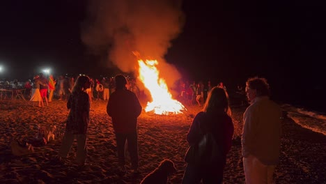 Big-crowd-at-the-traditional-bonfire-festival-at-the-beach-at-the-San-Juan-celebration-in-Marbella-Spain,-people-dancing-and-enjoying-a-fun-party-during-summer,-big-burning-fire-and-flames,-4K-shot