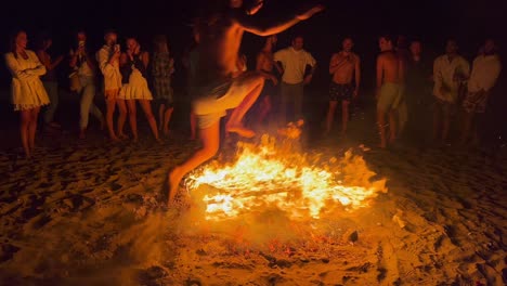 People-jumping-over-a-traditional-bonfire-at-the-San-Juan-festival-at-the-beach-in-Marbella-Spain,-friends-and-family-enjoying-a-fun-party,-jumping-over-a-big-burning-fire-and-hot-flames,-4K-shot